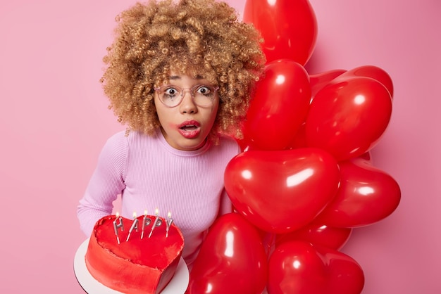 Shocked astonished European woman with curly hair stares impressed at camera cannot cannot believe in amazing news celebrates Valentines Day poses with tasty cake and bunch of heart balloons