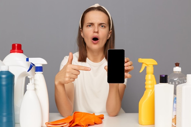 Shocked astonished surprised woman posing at workplace with cleaning detergents isolated over gray background pointing at blank display empty space for advertisement