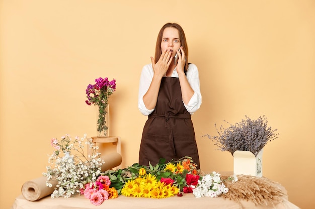 Shocked astonished woman entrepreneur wearing brown apron standing in own floral store and talking on smartphone with client posing isolated over beige background