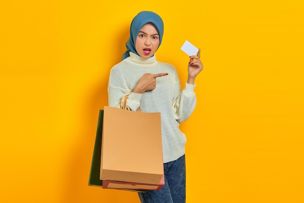 Shocked Beautiful Asian woman in white sweater holding shopping bag and pointing finger at credit card isolated over yellow background