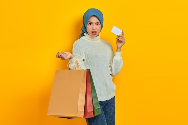 Shocked beautiful Asian woman in white sweater holding shopping bags and credit card isolated over yellow background