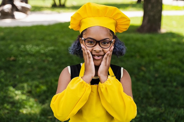 Shocked black african child cook girl in chefs hat and yellow apron uniform hold cheeks and surprise Creative advertising for cafe or restaurant