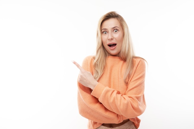 Shocked blond girl looks surprised on white studio background points finger at copy space