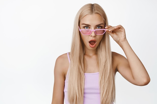 Shocked blond girl takes off sunglasses say wow and stare startled at camera standing in tank top against white background