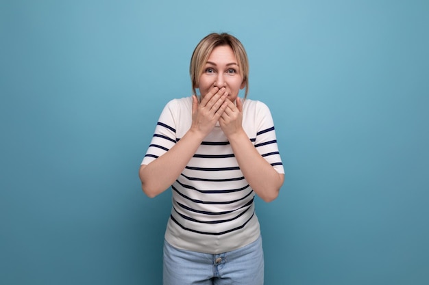 Shocked blond young woman in a striped sweater covered her mouth with her hand in surprise on a blue