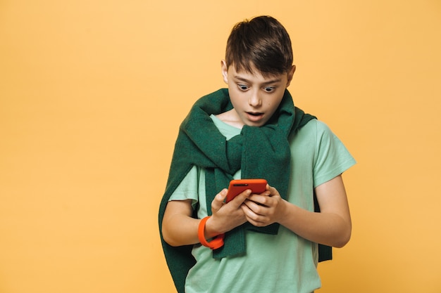 Shocked boy dressed in a green t-shirt and sweater tied up over his neck, holds his smartphone, looks at it with wide open eyes, amazed by received message. Education concept.