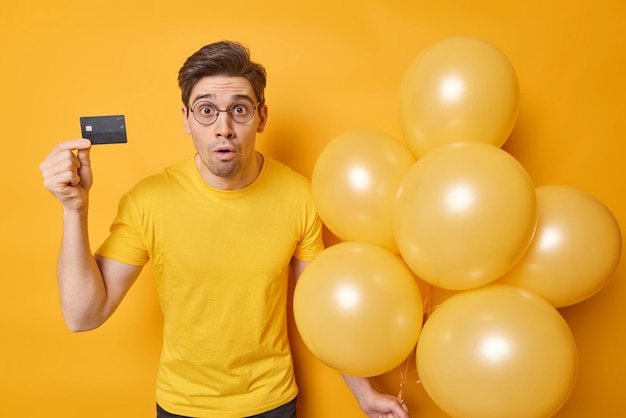 Shocked brunet man feels impressed dressed in casual t shirt holds credit card and bunch of inflated balloons prepares for festive event isolated over yellow background Festive occasion concept