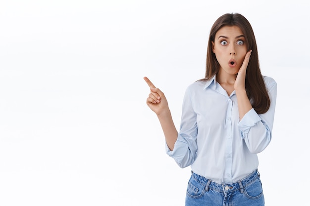 Shocked brunette woman gossiping with coalegues, gasping and drop jaw from rumor and shocking information, pointing finger upper left corner, hear interesting news, stand white wall