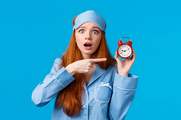 Shocked and concerned, worried cute redhead girl pointing at alarm clock with frustrated, nervous expression, being late, dont know how set up right time, standing blue wall in nightwear