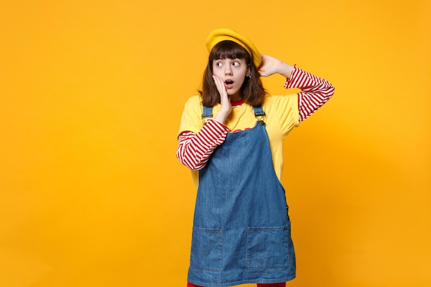 Shocked confused girl teenager in french beret, denim sundress keeping mouth open, putting hand on cheek, looking aside isolated on yellow wall background. People sincere emotions, lifestyle concept.