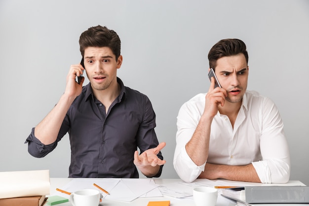 Shocked confused two young business men coworking talking by mobile phones.