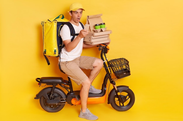 Shocked courier on electric scooter with thermo backpack delivering pizza and coffee to go holding cell phone looking at screen with open mouth isolated over yellow background