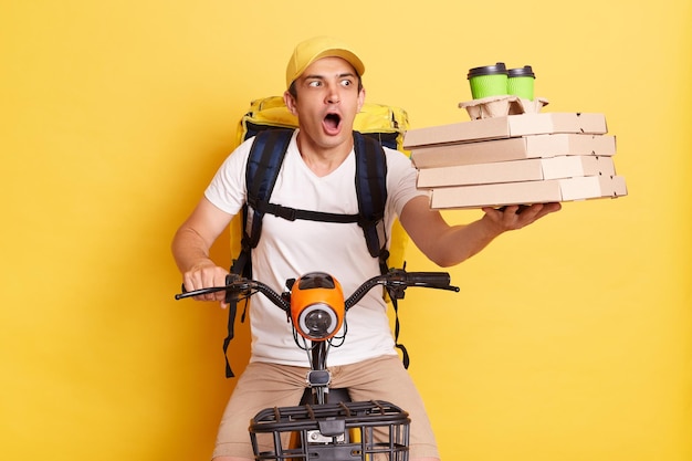 Shocked courier man holding pizza boxes and takeaway coffee cups delivering order on bike with big backpack isolated over yellow background looking away with open mouth wrong order