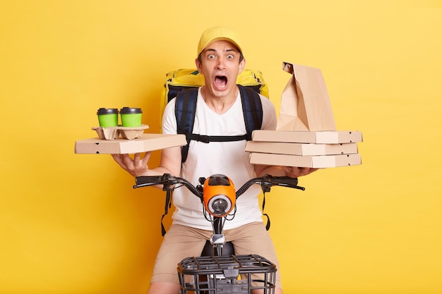 Shocked courier wearing cap and white t shirt holding pizza boxes and coffee riding bicycle looking at camera with open mouth and big eyes being scared isolated on yellow background