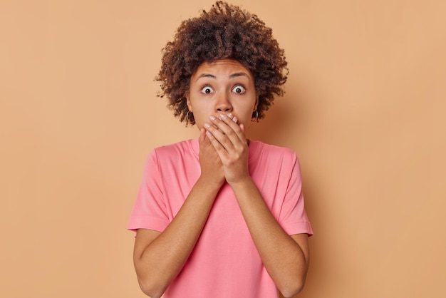 Shocked curly haired young woman covers mouth with hands stares scared at camera cannot believe in something horrible dressed in casual pink t shirt isolated over brown background Omg concept