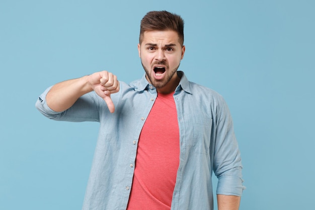 Shocked dissatisfied young bearded guy in casual shirt posing isolated on pastel blue background studio portrait. People sincere emotions lifestyle concept. Mock up copy space. Showing thumb down.