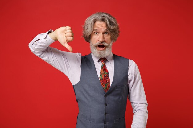 Shocked elderly gray-haired mustache bearded man in classic shirt vest and colorful tie isolated on red background, studio portrait. People lifestyle concept. Mock up copy space. Showing thumb down.