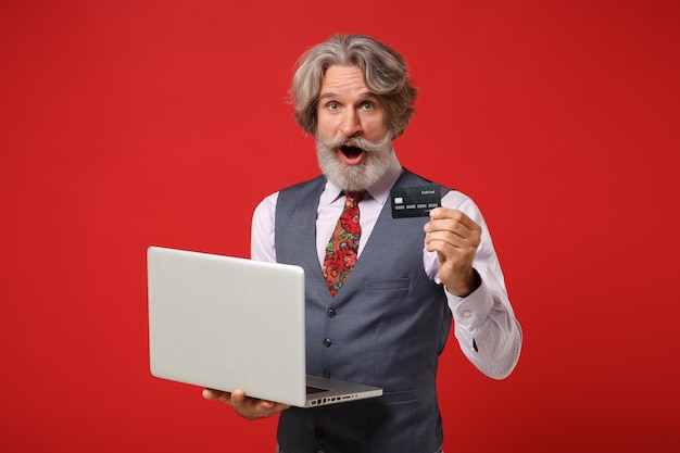 Shocked elderly gray-haired mustache bearded man in classic shirt vest tie posing isolated on red background. People lifestyle concept. Mock up copy space. Hold laptop pc computer, credit bank card.