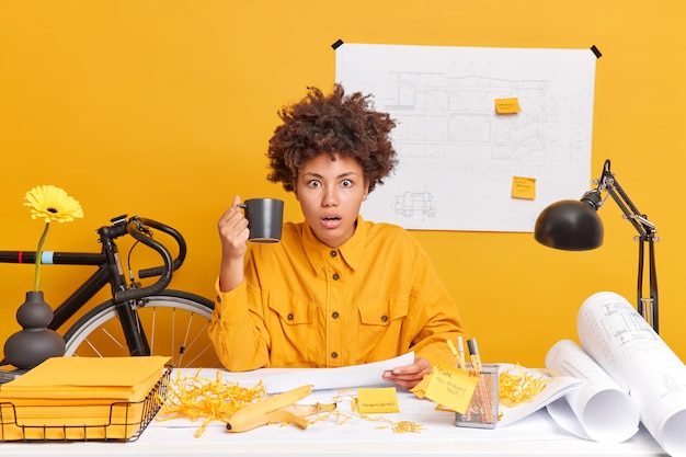 Shocked ethnic female architect works on detailed construction blueprints has coffee break poses at dekstop stares impressed, prepares project wears yellow shirt holds cup of beverage