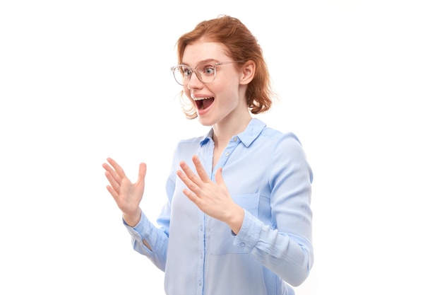 Shocked face redhaired girl in business shirt looks surprised with open mouth spreads hands on white studio background