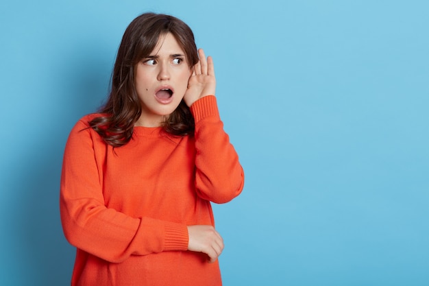 Shocked female wearing orange casual sweater posing with palm near the ear, trying to hear something, isolated over blue wall.