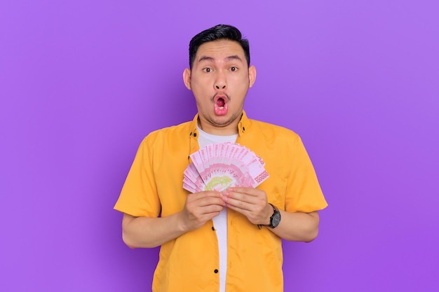 Shocked handsome young Asian man holding a fan of money banknotes isolated on purple background