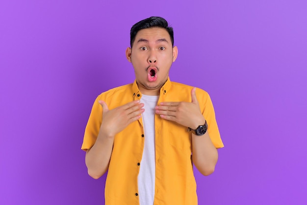 Shocked handsome young Asian man keeps hands on chest with omg expression isolated on purple background