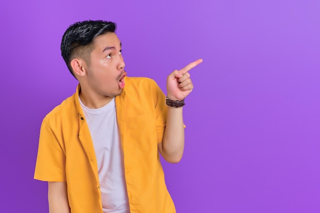 Shocked handsome young Asian man pointing fingers aside at copy space isolated on purple background