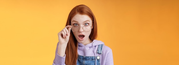 Photo shocked impressed redhead coworker hearing last intimate rumors drop jaw staring full disbelief camera confused touch glasses seem unsure truth lie standing orange background astonished