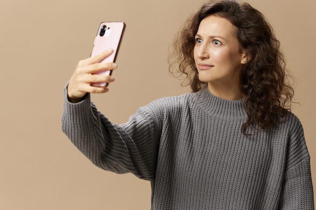 Shocked irritated curly lady in gray casual sweater do video call with phone get bad news posing isolated on over beige background Social media network distance communication concept Copy space