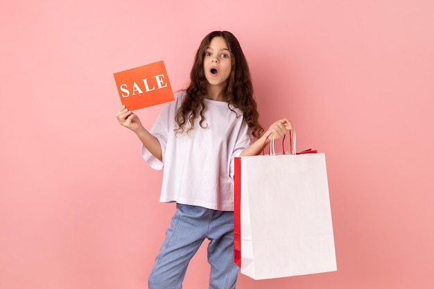 Shocked little girl holding shopping bags and sale card astonished with big holidays discounts