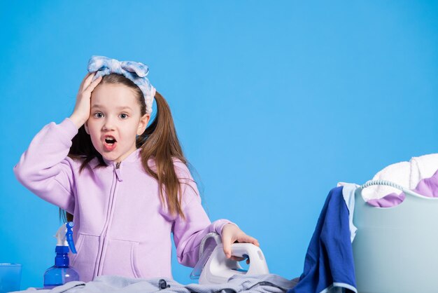 A shocked little girl sees how much ironing awaits her The surprised child holds the iron on the ironing board the daughter helps mother with the housework catches head tired