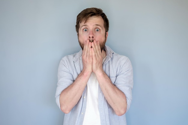 Shocked man in casual clothes puts hands over mouth in fear and looks at the camera with big eyes