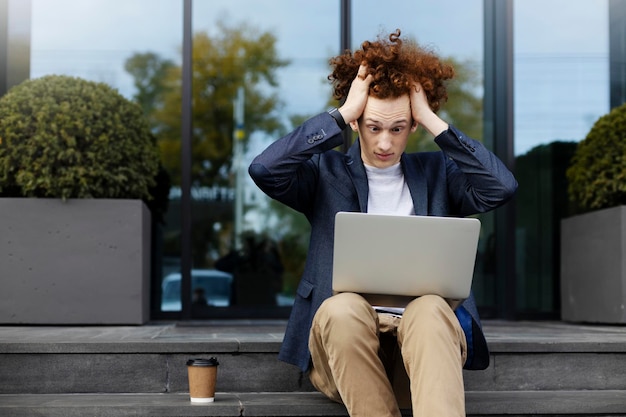 Shocked man using laptop holding his hair he has a problem Surprised man sitting on stairs outdoors