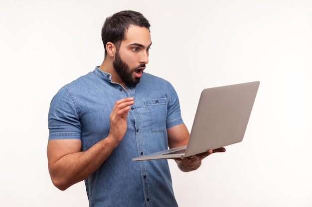 Shocked man with beard looking at laptop display with opened mouth and big eyes surprised with operation system error Indoor studio shot isolated on white background