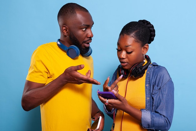 Shocked man and woman pointing at mobile phone screen, finding unexpected news on social media. Surprised couple having fun with headphones and modern smartphone to browse internet.