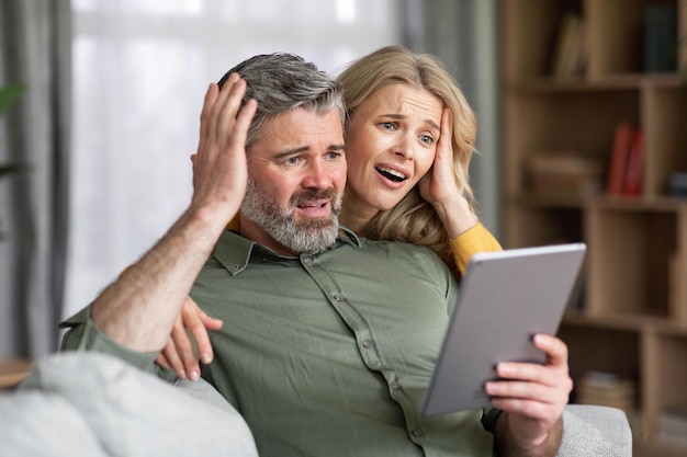 Shocked middle aged spouses looking at digital tablet screen at home