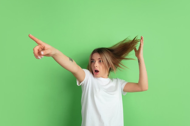 Shocked pointing up. Caucasian young woman's portrait isolated on green studio wall