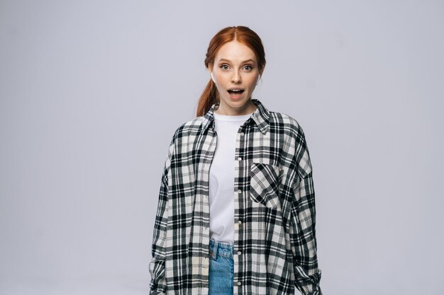 Shocked redhaired young woman wearing wireless earphones looking at camera on isolated white background Pretty redhead lady model emotionally showing facial expressions copy space