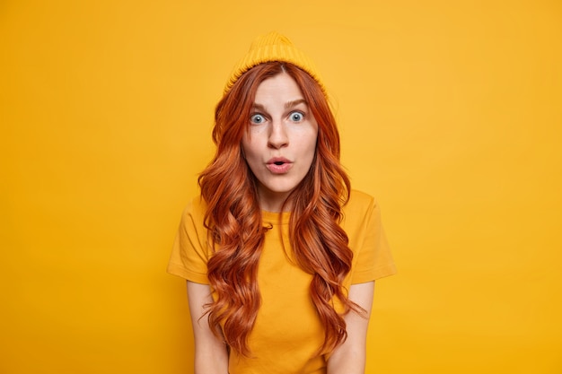 Shocked redhead girl stares speechless, has starled face expression opens mouth from amazement wears hat casual t shirt 