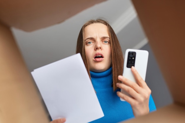 Shocked sad woman received parcel with wrong or damages items holding waybill in hands and mobile phone calling to service looking inside of box with astonished expression