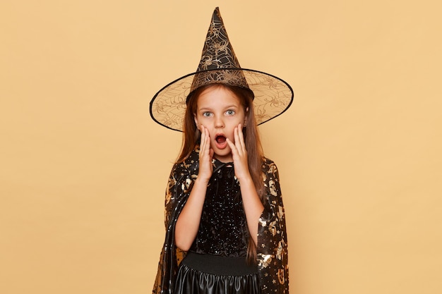 Shocked scared little girl wearing witch costume and carnival cone hat isolated over beige background looking at camera with open mouth spooky surprise at halloween party