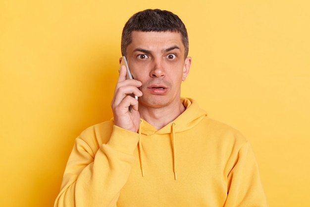 Shocked scared man wearing casual hoodie talking on smart phone hearing surprised news looking at camera with big eyes posing isolated over yellow background