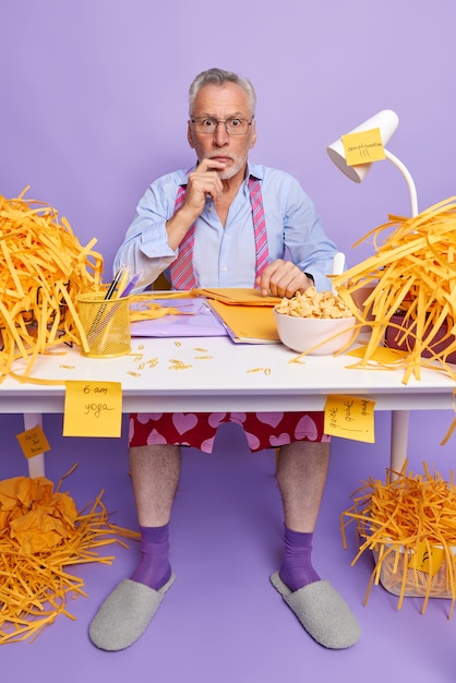 Shocked senior man work from home sits at white desk with folders paperclips bowl of cornflakes cut paper weras transparent glasses formal shirt tie around neck socks and slippers purple wall