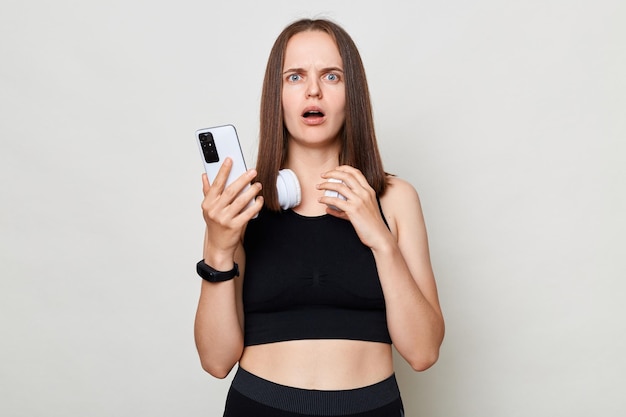 Shocked slim woman with headphones wearing sportswear posing against gray background reading new in social network berowsing internet