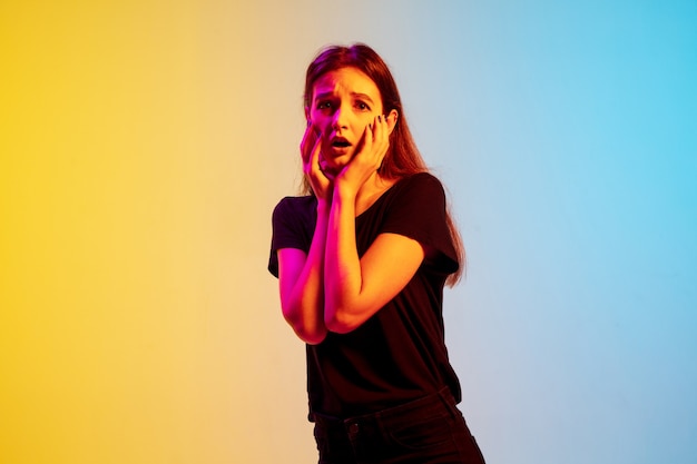 Shocked, stressed. Young caucasian woman's portrait on gradient blue-yellow studio background in neon light. Concept of youth, human emotions, facial expression, sales, ad. Beautiful brunette model.