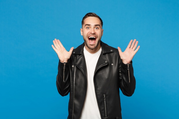 Shocked stylish young bearded man in black leather jacket white t-shirt looking camera isolated on blue wall background studio portrait. People sincere emotions lifestyle concept. Mock up copy space.