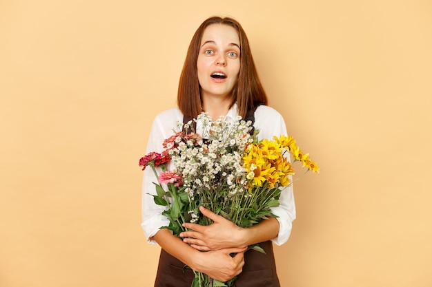 Shocked surprised amazed adult woman florist wearing brown apron holding bouquet standing isolated over beige background with beautiful flower composition