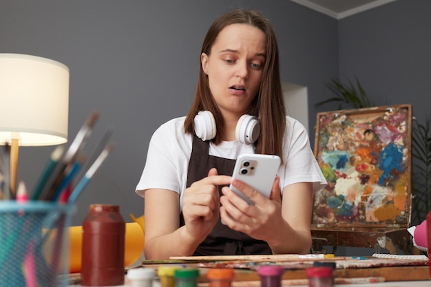 Photo shocked surprised brown haired woman artist drawing with paints and using mobile phone sitting in studio workshop looking at device screen with amazed facial expression