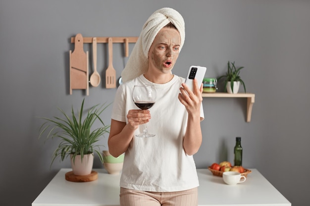 Shocked surprised woman with cosmetic mask in white towel wrapped around head drinking wine in kitchen holding smart phone in hand looking at screen with open mouth reading breaking news